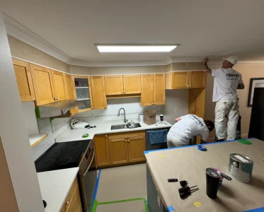 Kitchen under renovation with two workers, one on a ladder, updating cabinets and appliances.