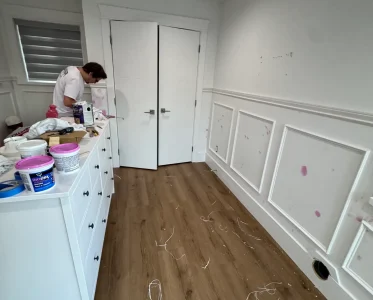 Person working on a project near a sink in a room with a wooden floor and scattered bottles.
